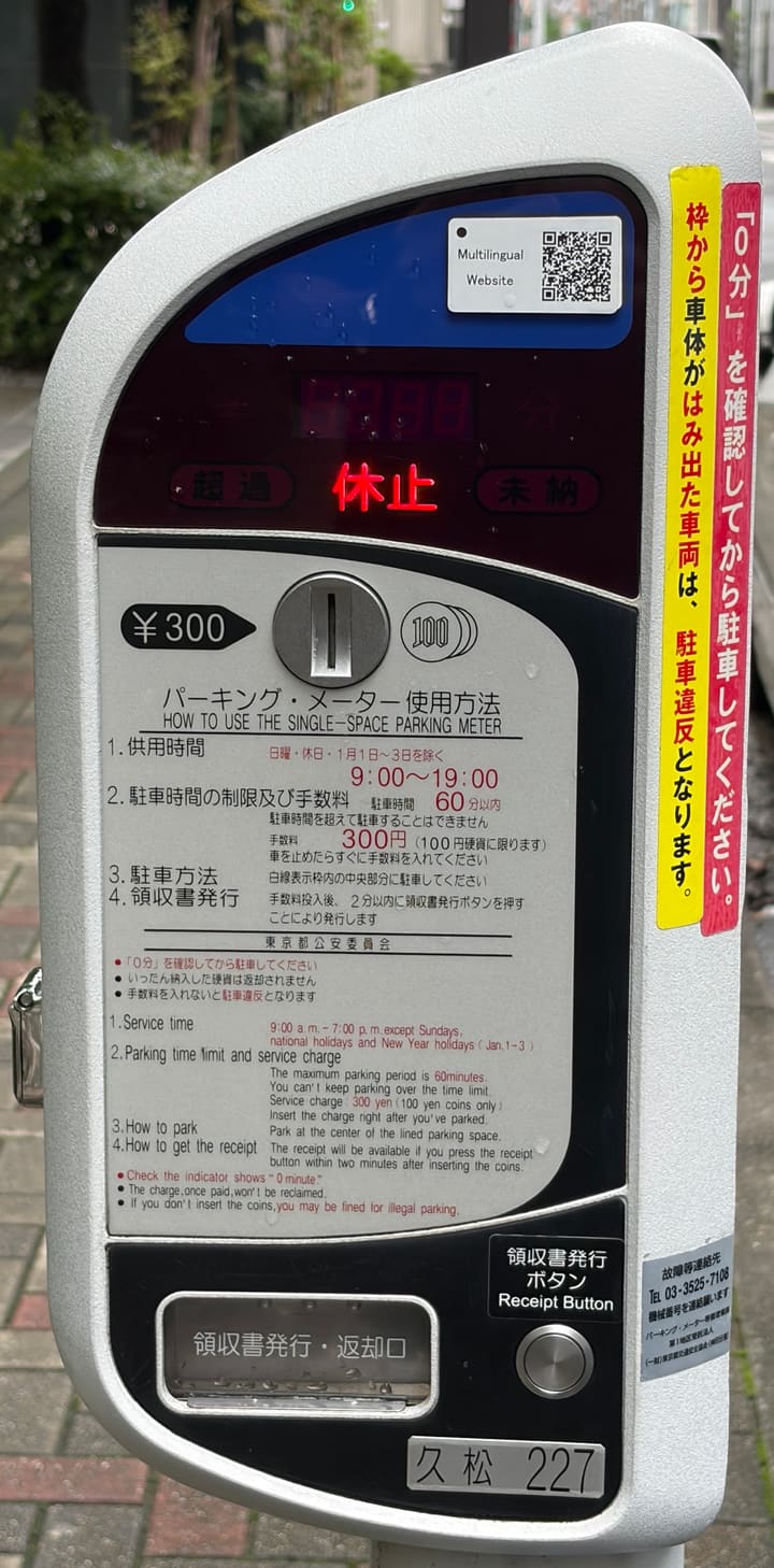 A Tokyo parking meter showing 休止 on its display, a 300 yen coin slot for 100-yen coins, bilingual instructions, a receipt button and a Multilingual Website QR code